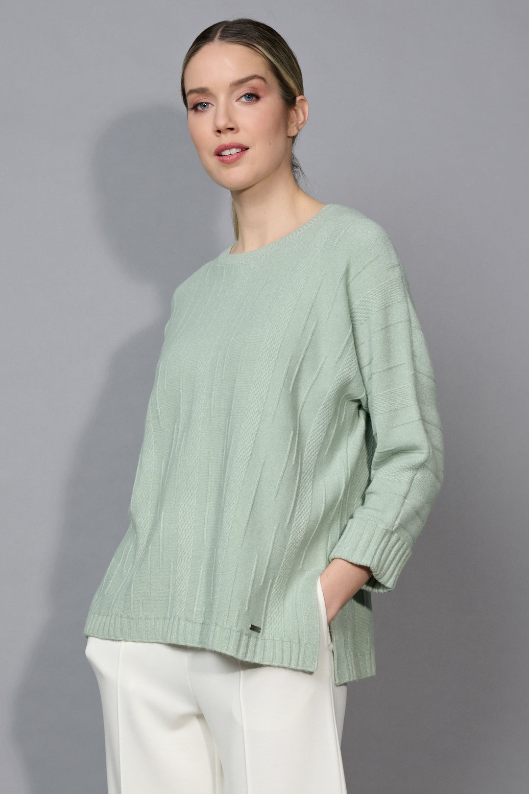 Woman wearing a light green sweater and white pants against a gray background