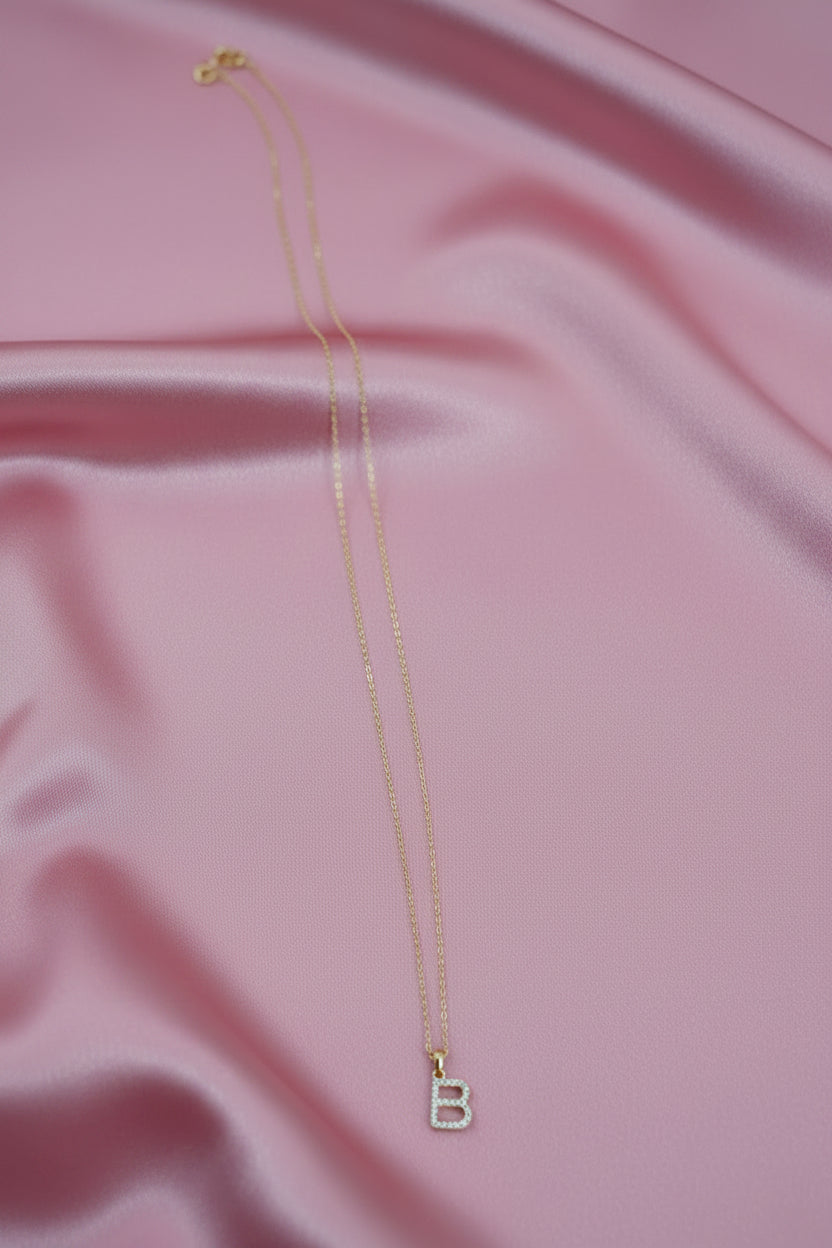A delicate gold chain necklace with a letter "B" cubic zirconia monogram charm, laid on pick stain piece of cloth.