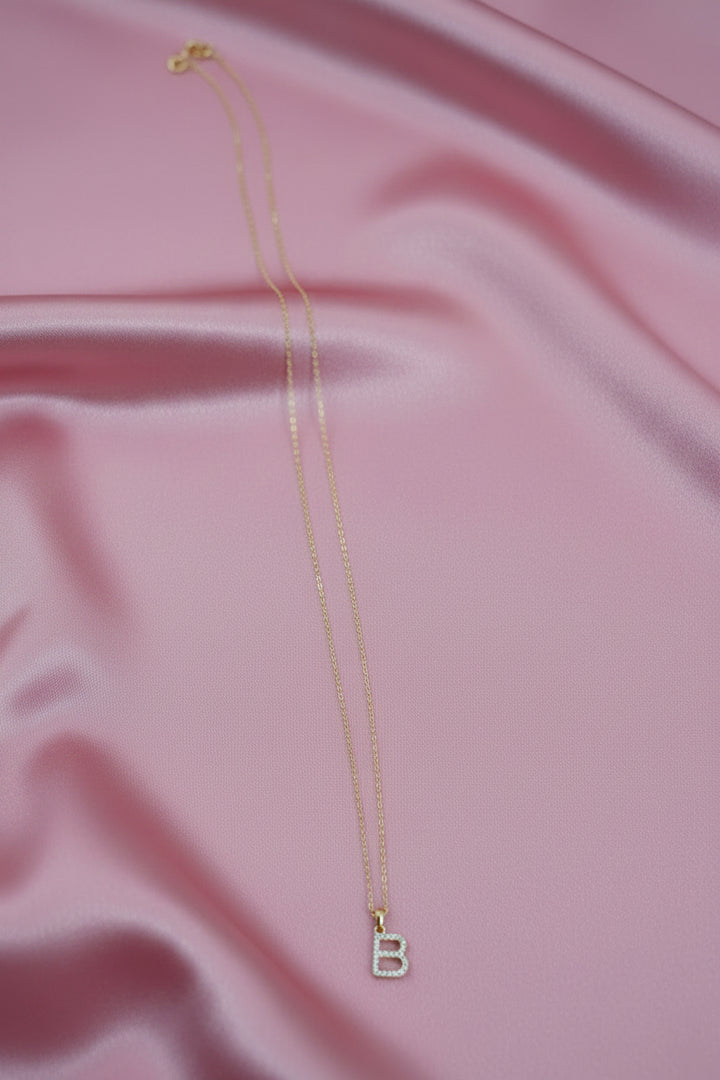 A delicate gold chain necklace with a letter "B" cubic zirconia monogram charm, laid on pick stain piece of cloth.