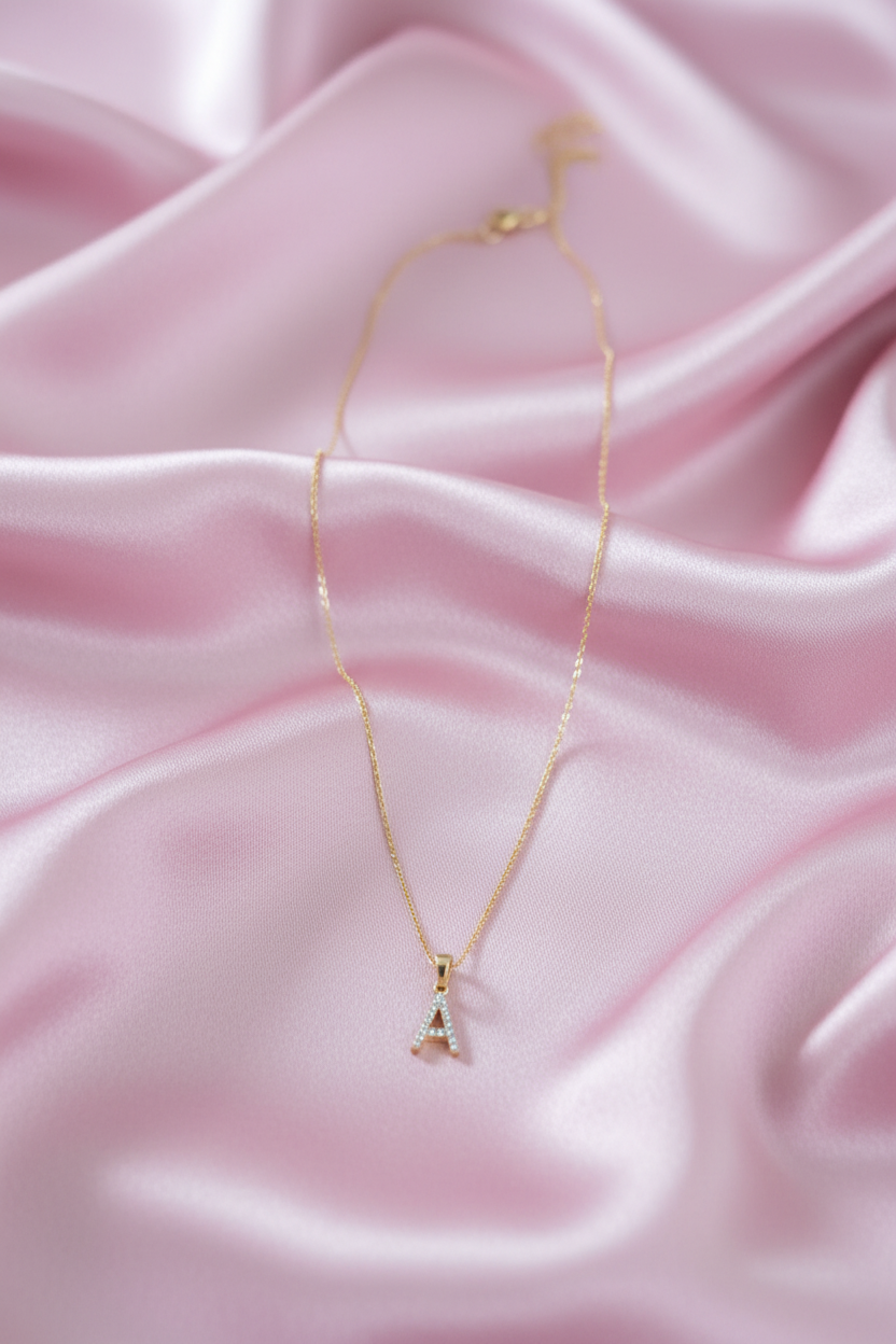 A delicate gold necklace with a letter 'A' initial pendant adorned with small clear stones, resting on draped pink satin fabric.