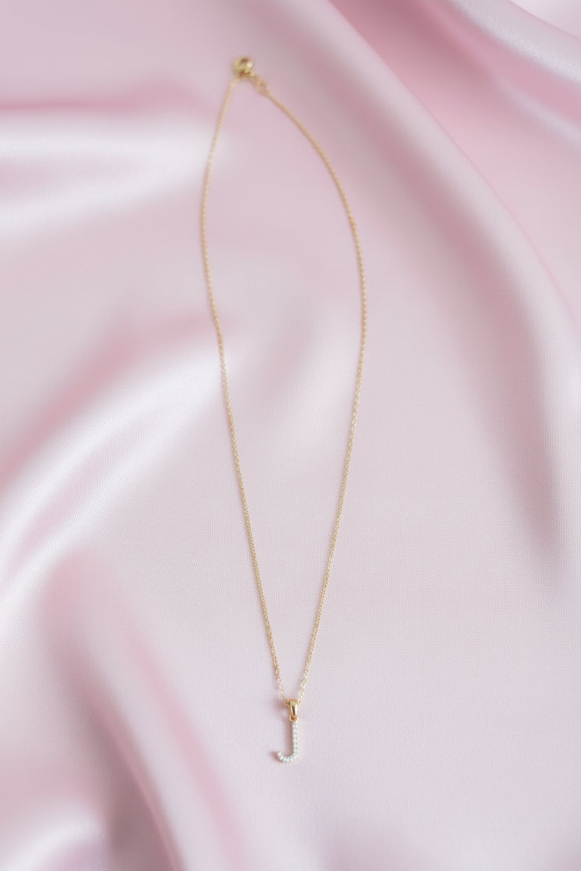 A delicate gold chain necklace with a small cubic zirconia stones J-shaped pendant, laid out on a pink satin cloth.