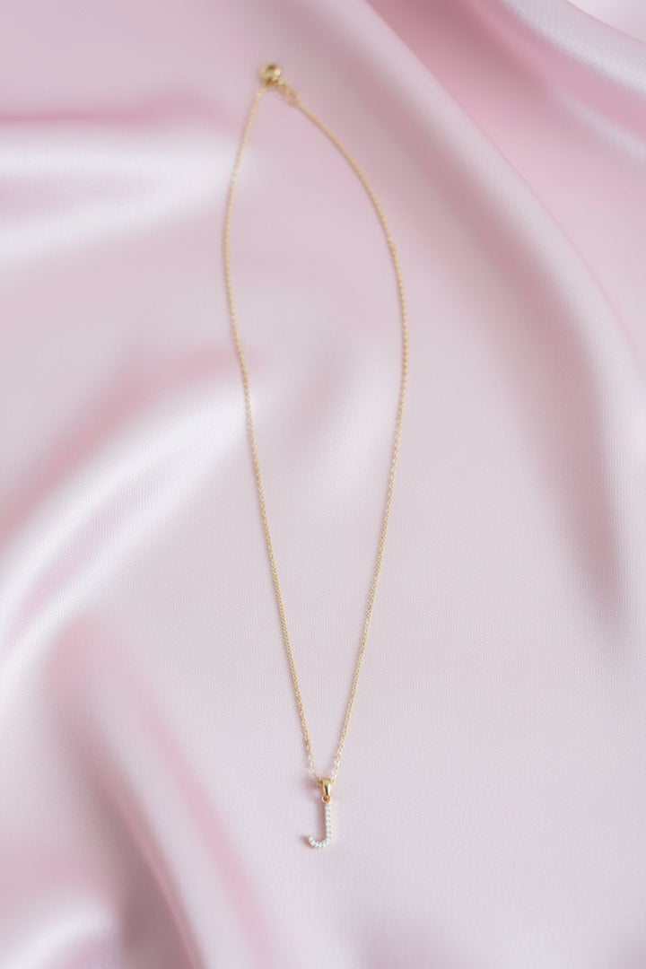 A delicate gold chain necklace with a small cubic zirconia stones J-shaped pendant, laid out on a pink satin cloth.
