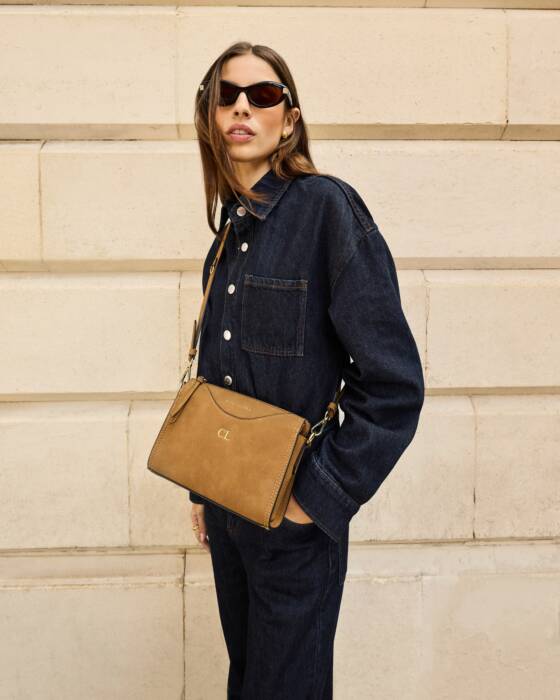 Woman wearing a dark denim jacket and holding a brown leather bag against a beige wall.