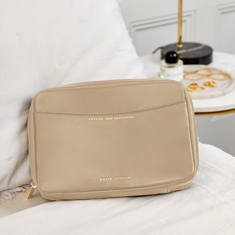 A beige cable organizer with a zip closure and a branded label on the front, placed on a white surface next to a pillow and a lamp.