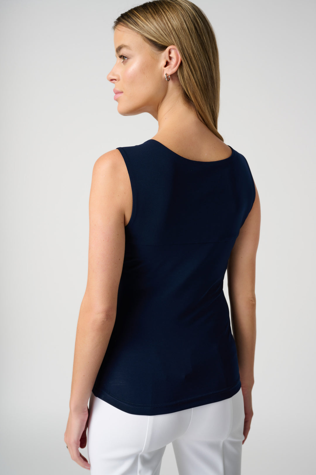 A back view of a woman wearing a navy sleeveless top and white pants on a light gray background