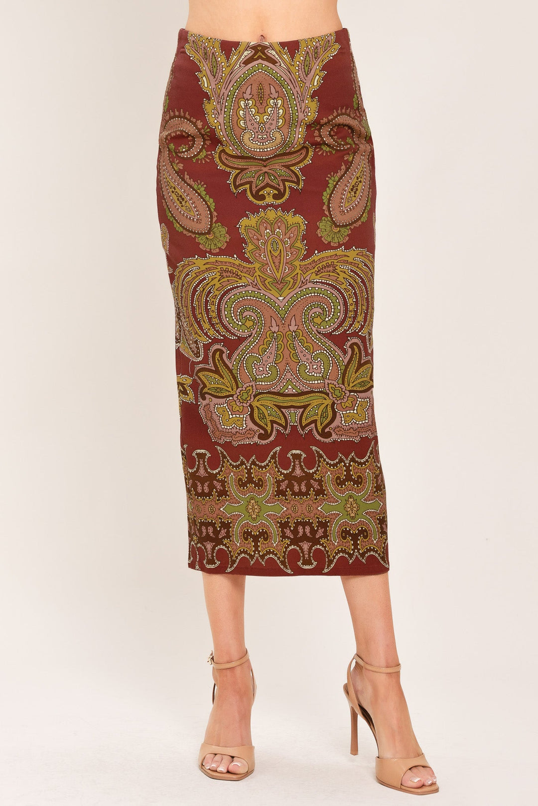 A woman models a knee-length fitted skirt with an ornate paisley pattern in rust red, gold, and olive green, paired with beige high-heeled sandals against a plain light background.