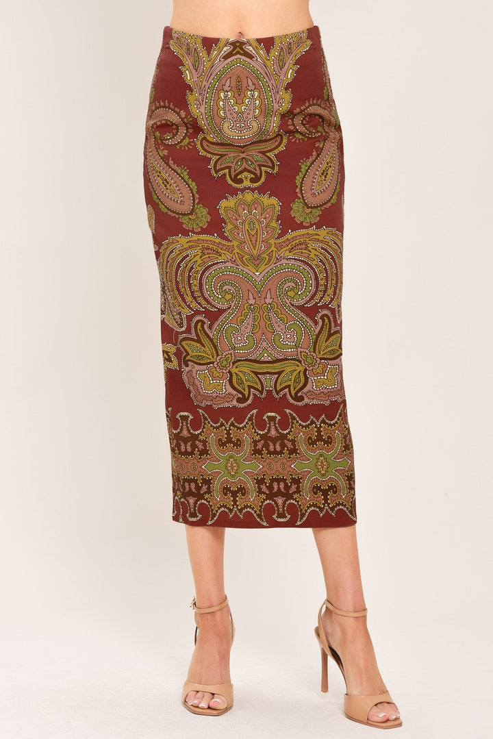 A woman models a knee-length fitted skirt with an ornate paisley pattern in rust red, gold, and olive green, paired with beige high-heeled sandals against a plain light background.