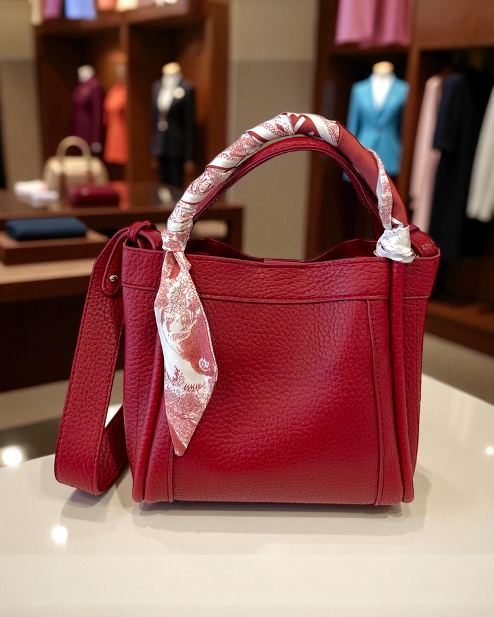 Red handbag with a decorative ribbon on a white surface, with a clothing store background.