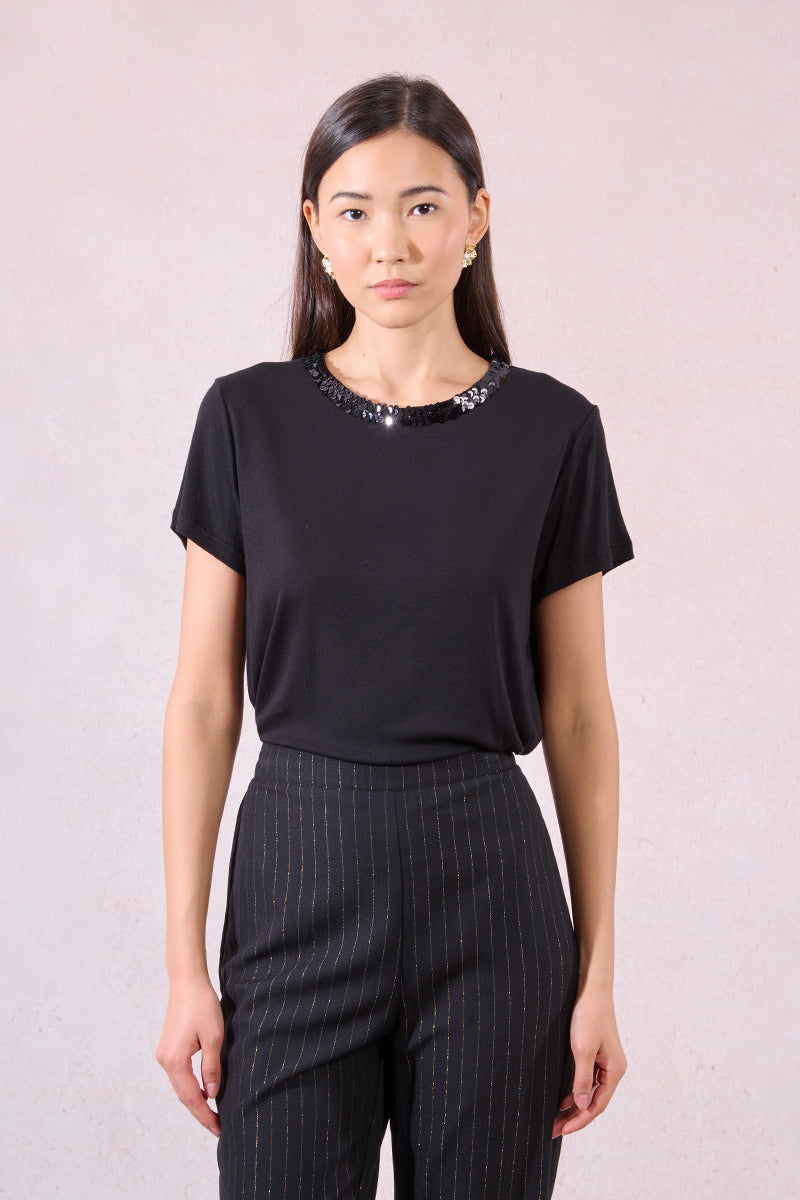 Woman wearing a black top with sequin details and black pinstripe pants against a light pink background