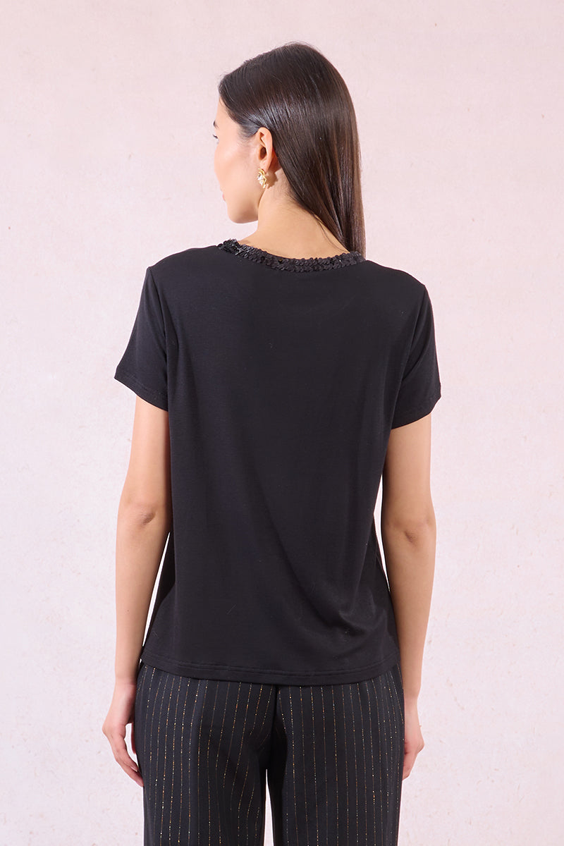 A back view of a woman wearing a black short-sleeve top with decorative trim on a light pink background
