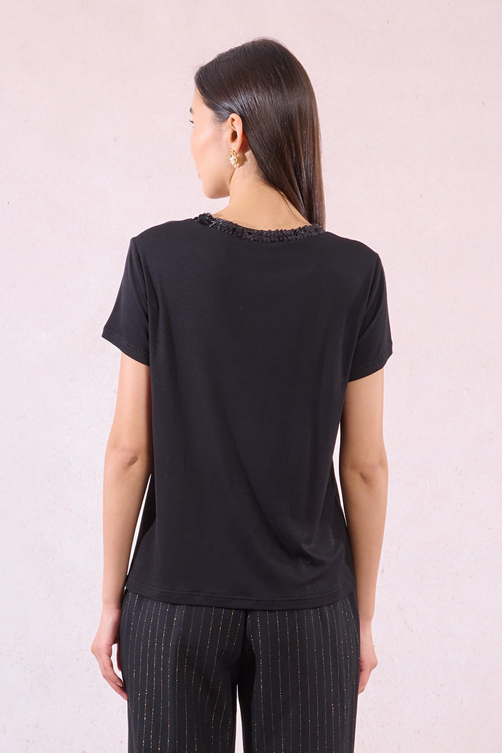 A back view of a woman wearing a black short-sleeve top with decorative trim on a light pink background